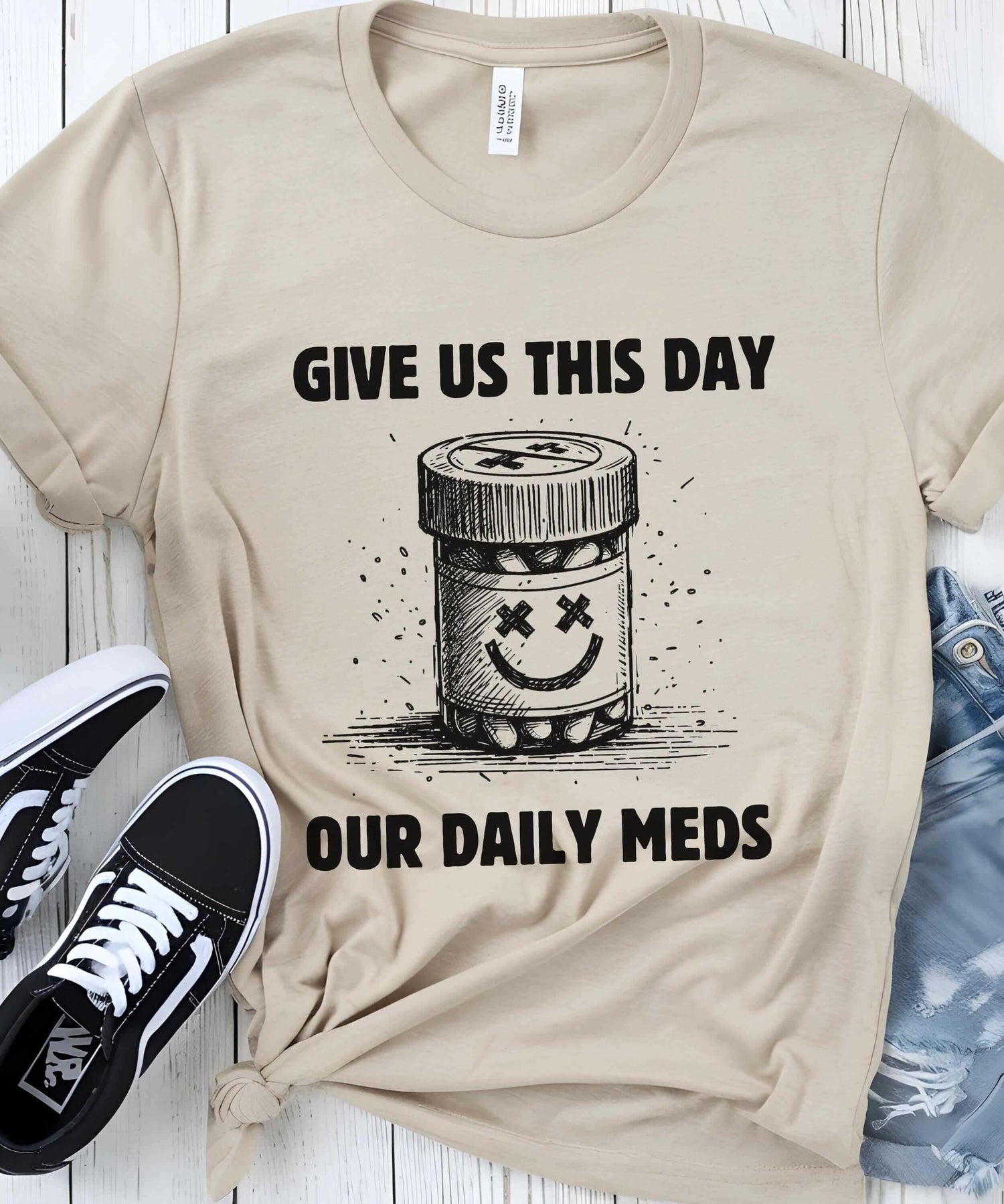 T-Shirt featuring a humorous design with a pill bottle and the text 'Give Us This Day Our Daily Meds'.